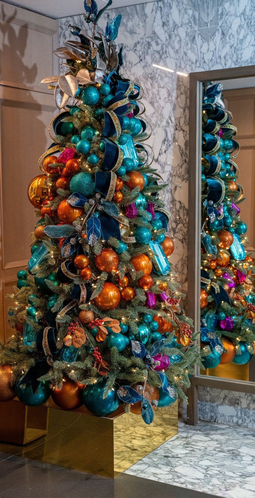 Large designer holiday tree in a marble hospitality lobby, styled in teal, copper, and gold with a mirrored gold base
